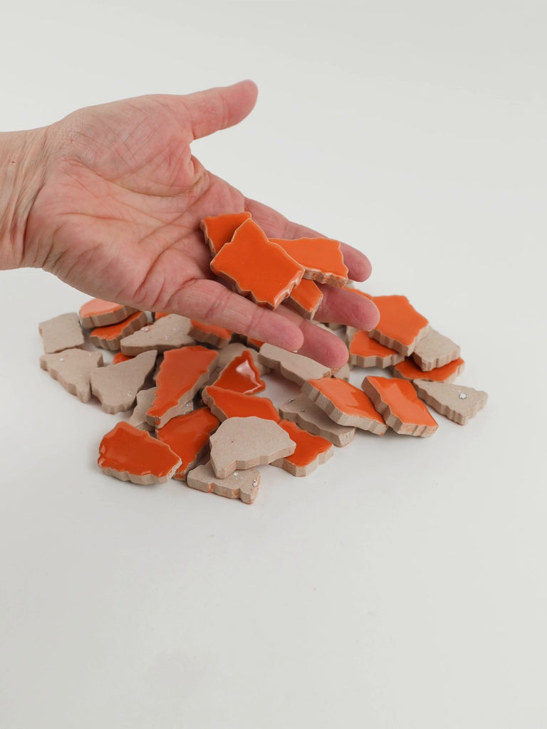 Video of Flip XL ceramic puzzle pieces in mandarin, opaque glazed porcelain, 20–60 mm, picked up from a pile by hand and dropped back to show their size, shapes and finish